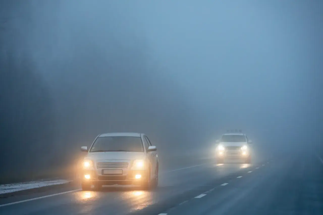 Deux voitures circulent sur une route brumeuse, leurs feux de brouillard avant allumés pour percer le brouillard épais.
