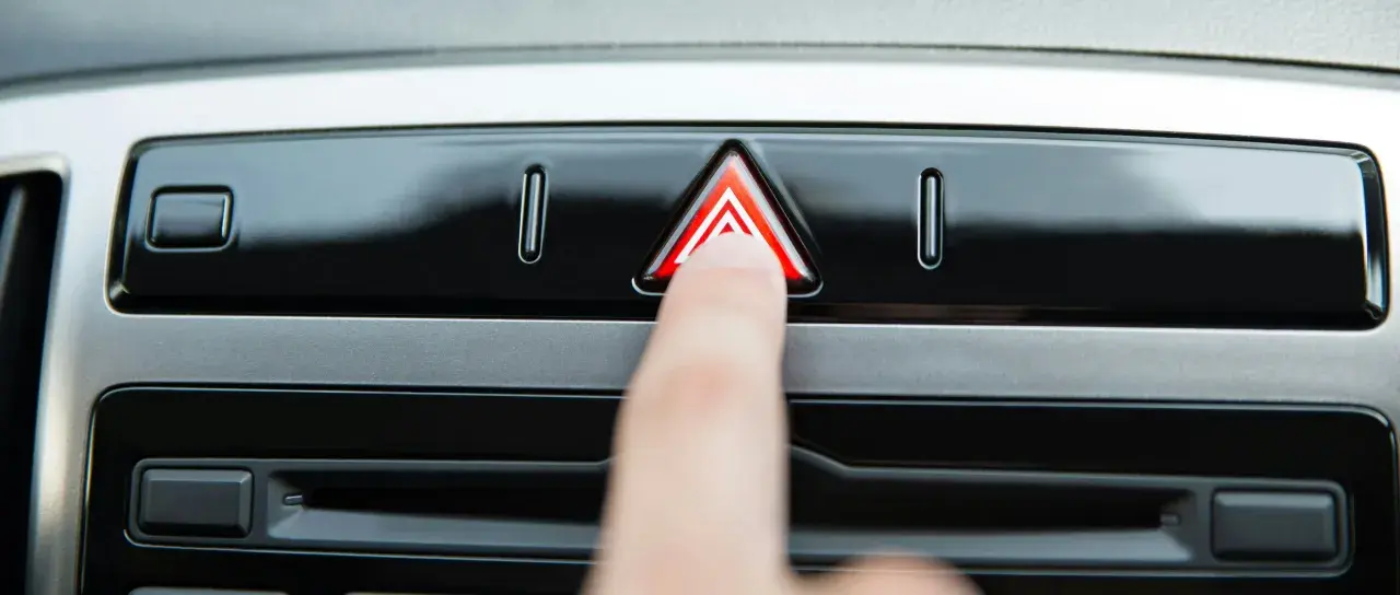 Un doigt appuie sur le bouton des feux de détresse, un triangle rouge lumineux sur le tableau de bord d'une voiture.
