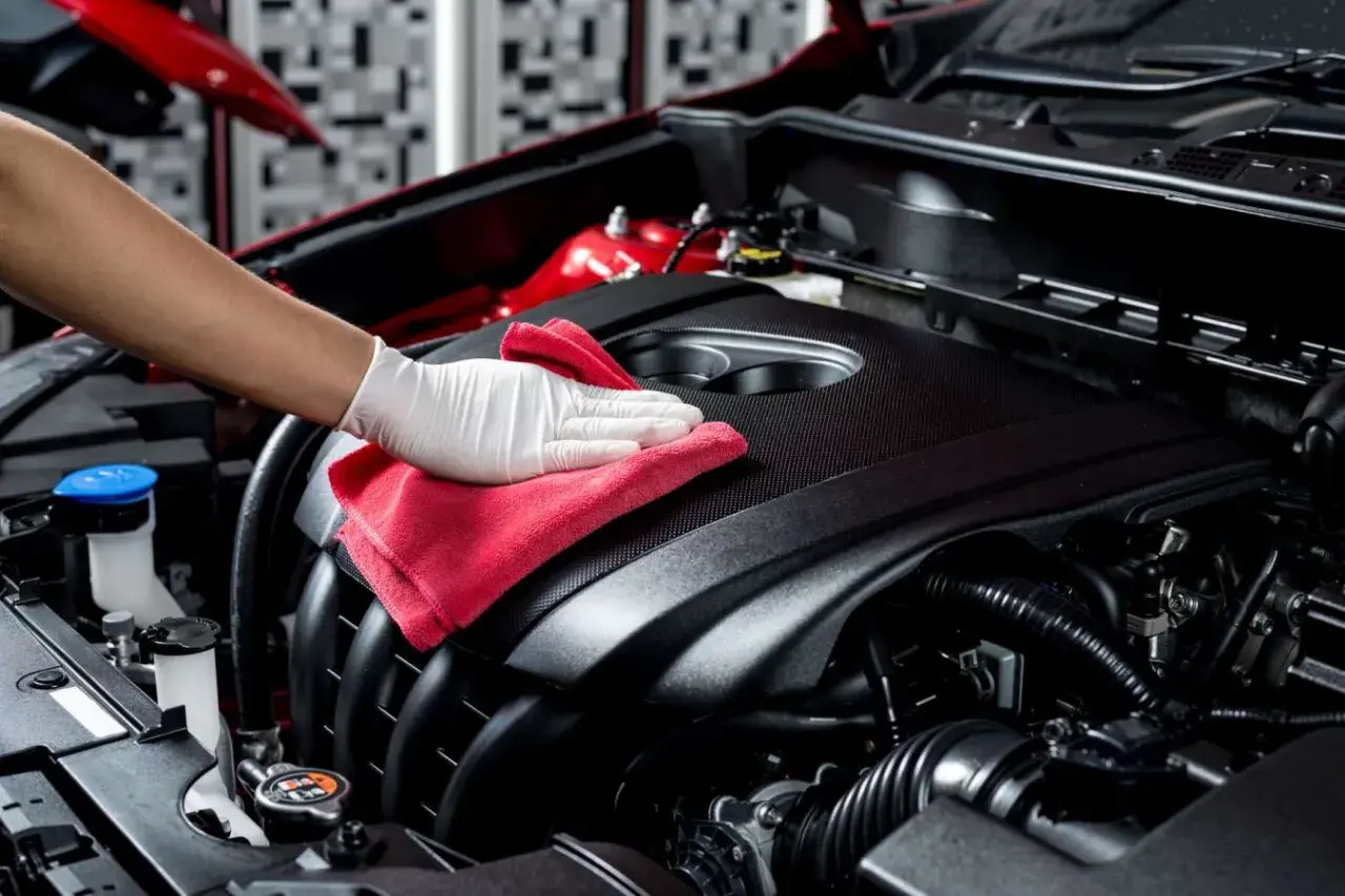 Main d'une personne portant un gant blanc, nettoyant le moteur d'une voiture avec un chiffon rouge.