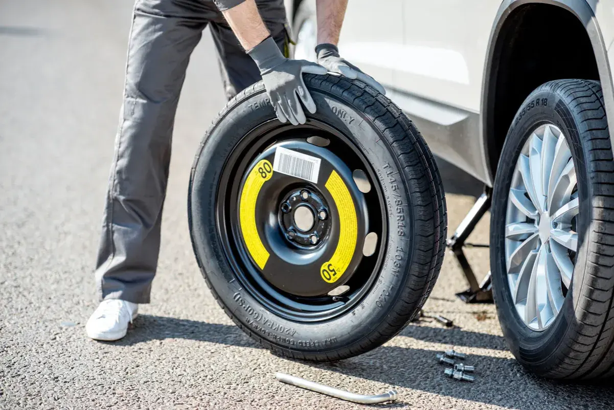 Un homme en gants gris tient une roue de secours pour changer un pneu. La voiture est sur un cric.
