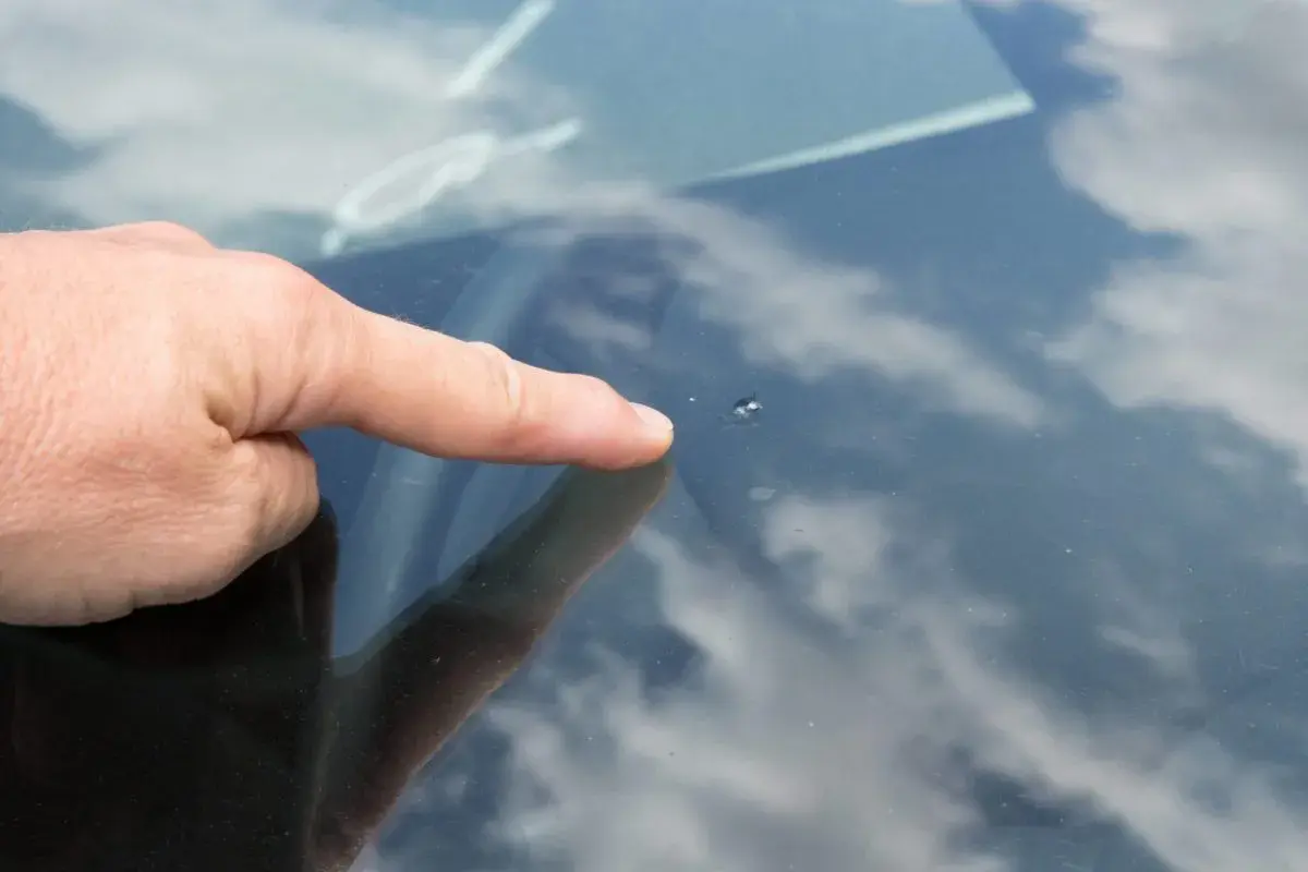 Un doigt pointe un petit impact pare brise sur le verre, reflétant le ciel nuageux.