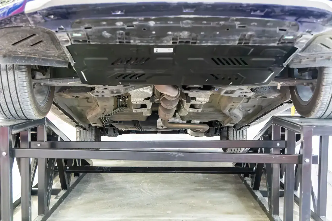 Vue du dessous d'une voiture sur des chandelles. Le système d'échappement est visible, ainsi qu'une plaque de protection. On dirait qu'il y a eu un choc sous la voiture.