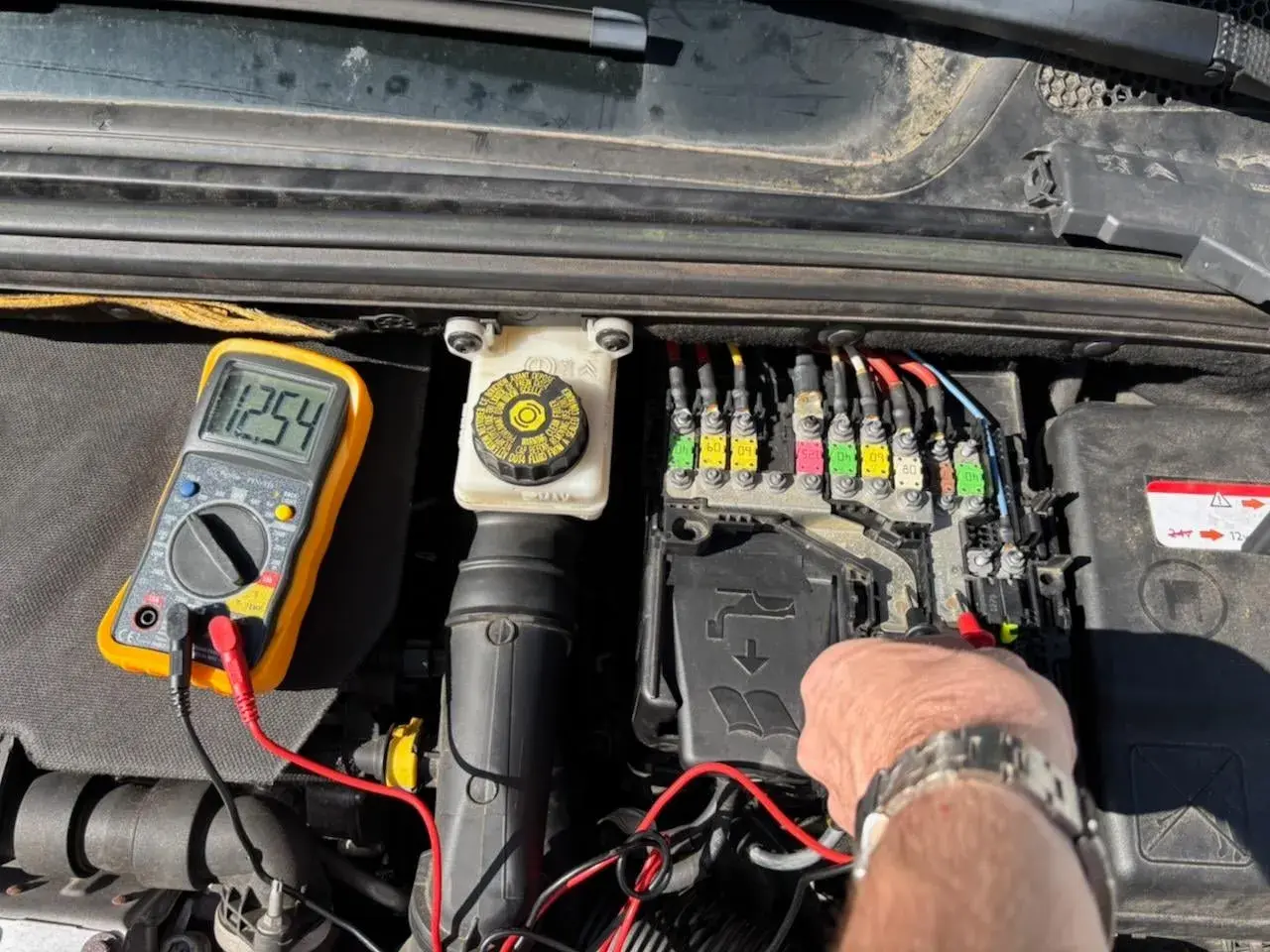 Diagnostic d'une C4 phase 2 : un multimètre affiche 12.54V près du boîtier de fusibles, indiquant une vérification avant de changer la batterie.