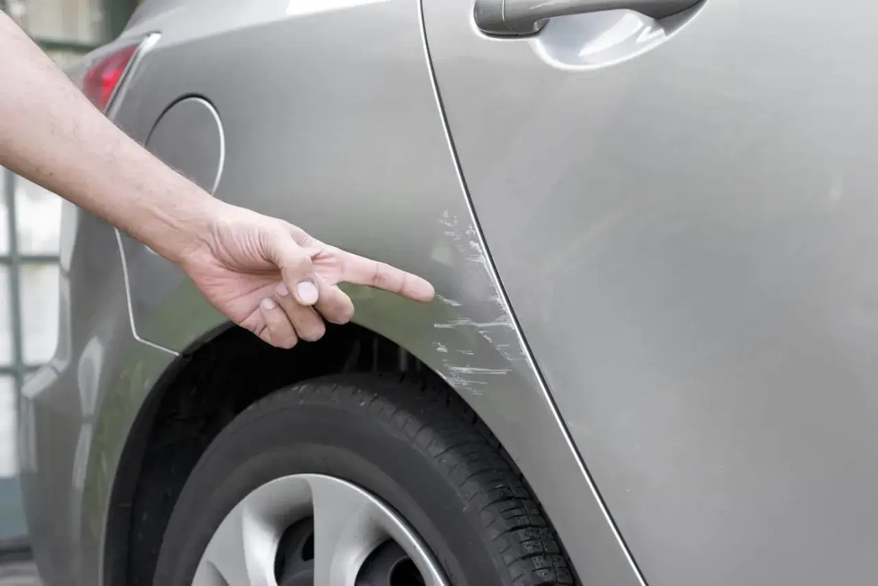 Doigt pointant des éraflures sur une voiture, résultat d'un frottement dans un parking.