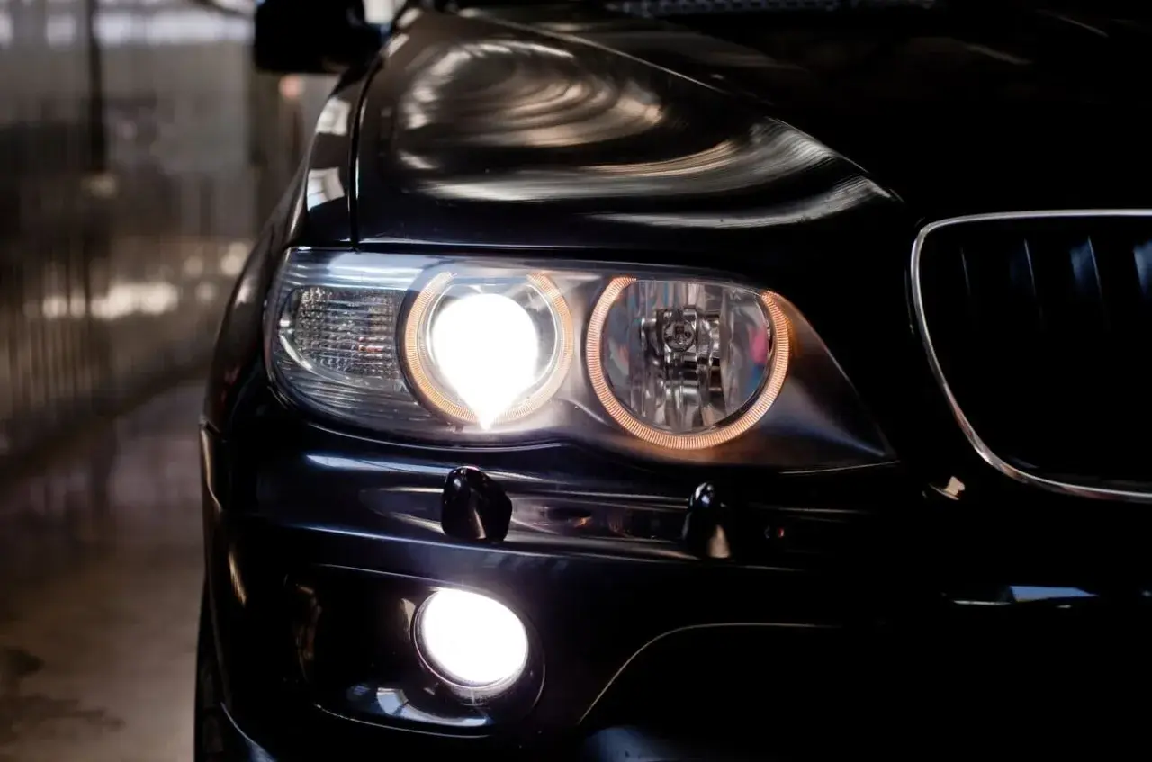 Gros plan sur l'avant d'une voiture noire, les feux de position allumés, créant une lumière intense et des reflets sur la carrosserie.