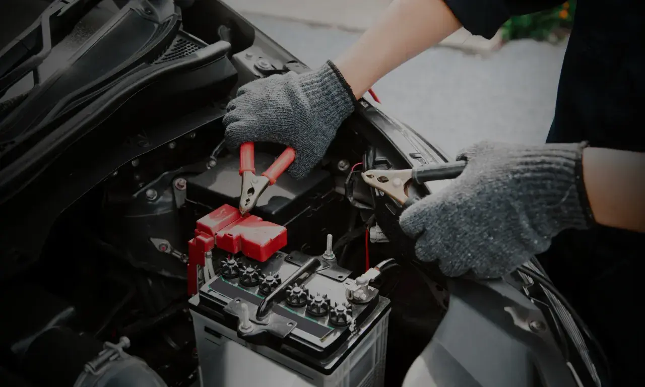 Mains de personne avec des gants gris connectant des câbles à une batterie de voiture. Un symptôme de batterie HS est souvent un démarrage difficile.