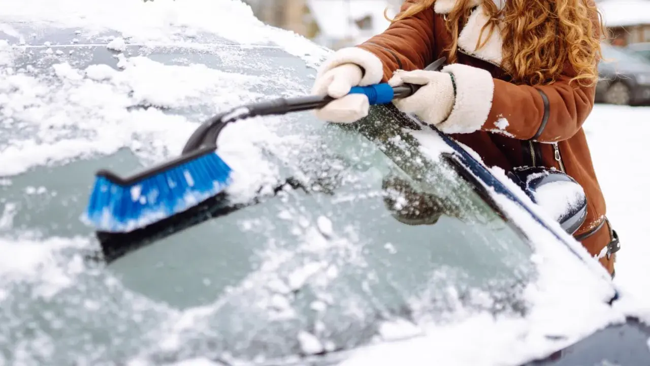 Une personne en manteau brun enlève la neige du pare-brise de sa voiture avec une brosse bleue. Faut il enlever la neige sur la voiture ? Oui, pour la sécurité !