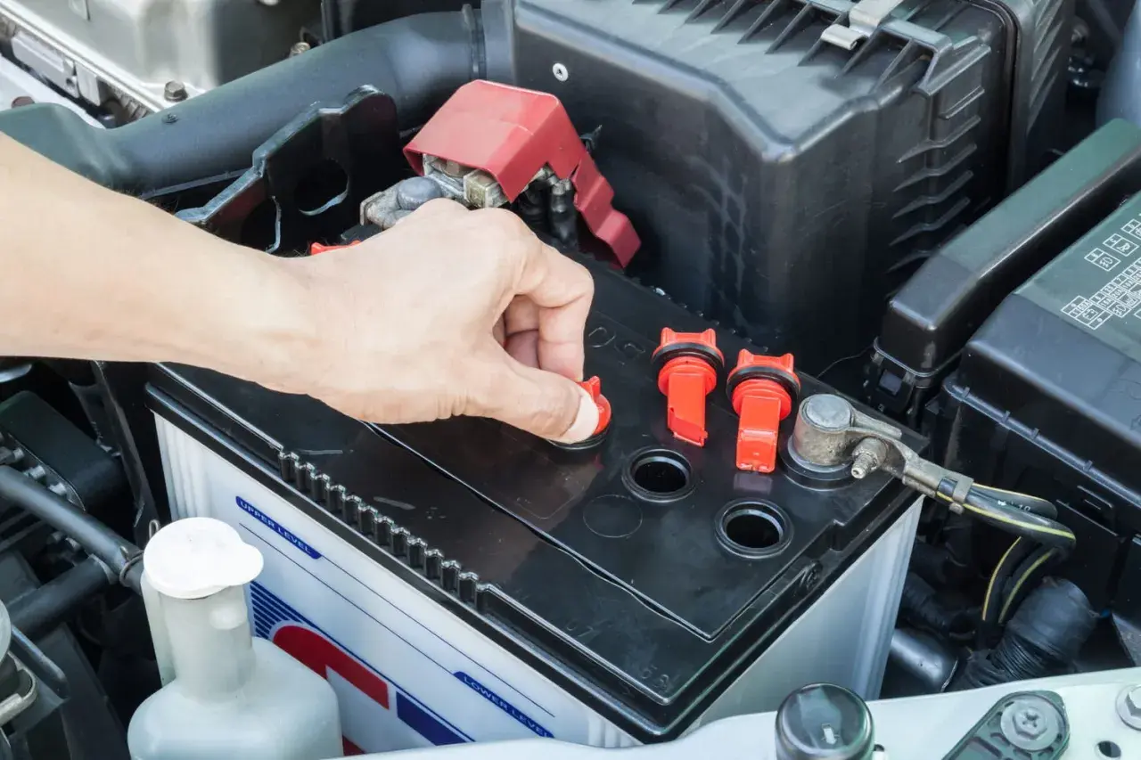Main d'une personne ouvrant un bouchon sur une batterie de voiture, peut-être pour vérifier le niveau d'eau pour batterie.