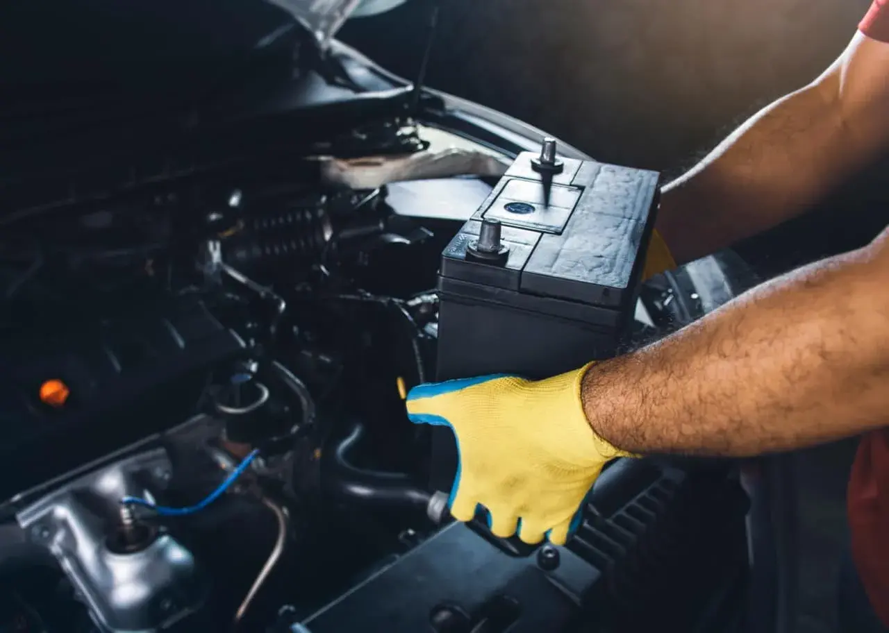 Mains pour gants jaunes tenant une batterie de voiture pour la débrancher. L'intérieur du compartiment moteur est visible.