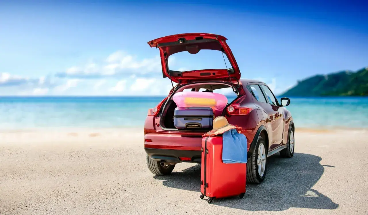 Voiture rouge prête pour le départ en vacances, coffre ouvert rempli de bagages, valise rouge et chapeau de paille sur le sable, mer bleue en arrière-plan.