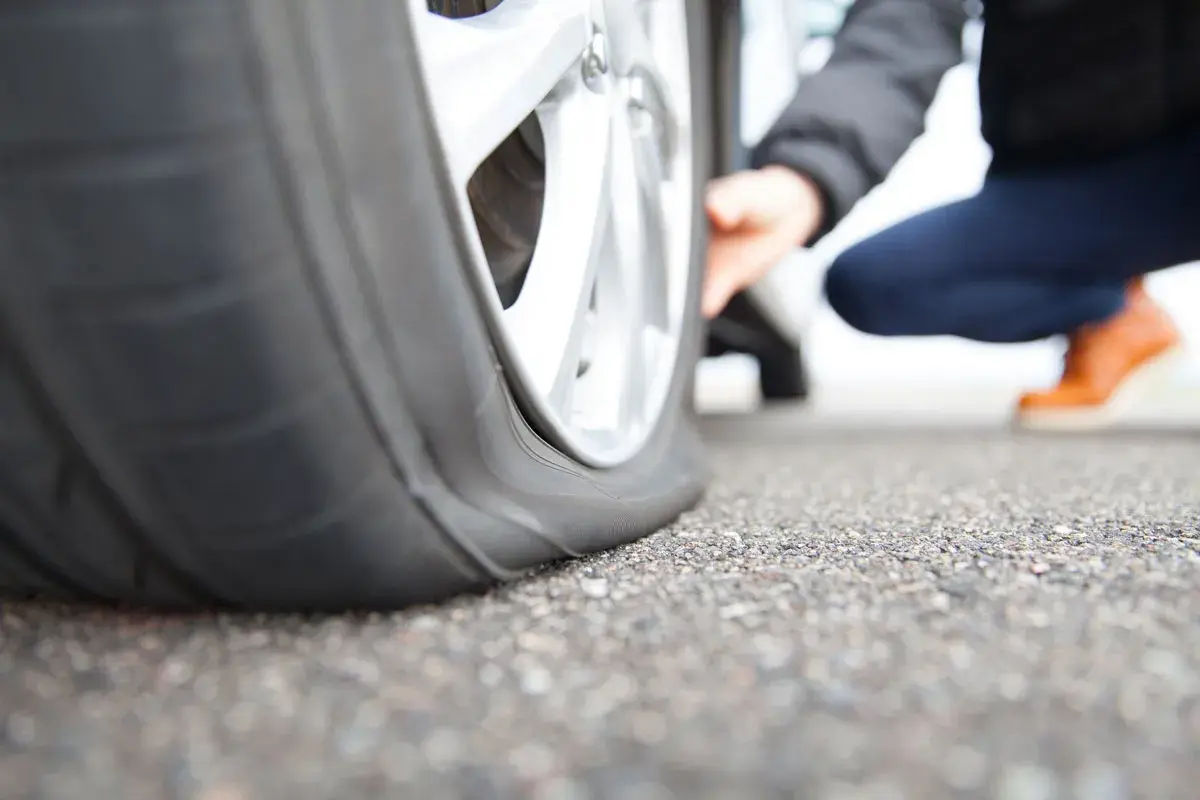 Gros plan sur un pneu crevé, une personne accroupie vérifie les dégâts. La route est asphaltée.