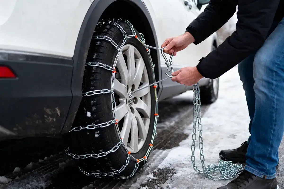 Un homme attache des chaînes neige à un pneu de voiture sur une route enneigée. Il est temps de savoir comment mettre des chaînes neige pour plus de sécurité.