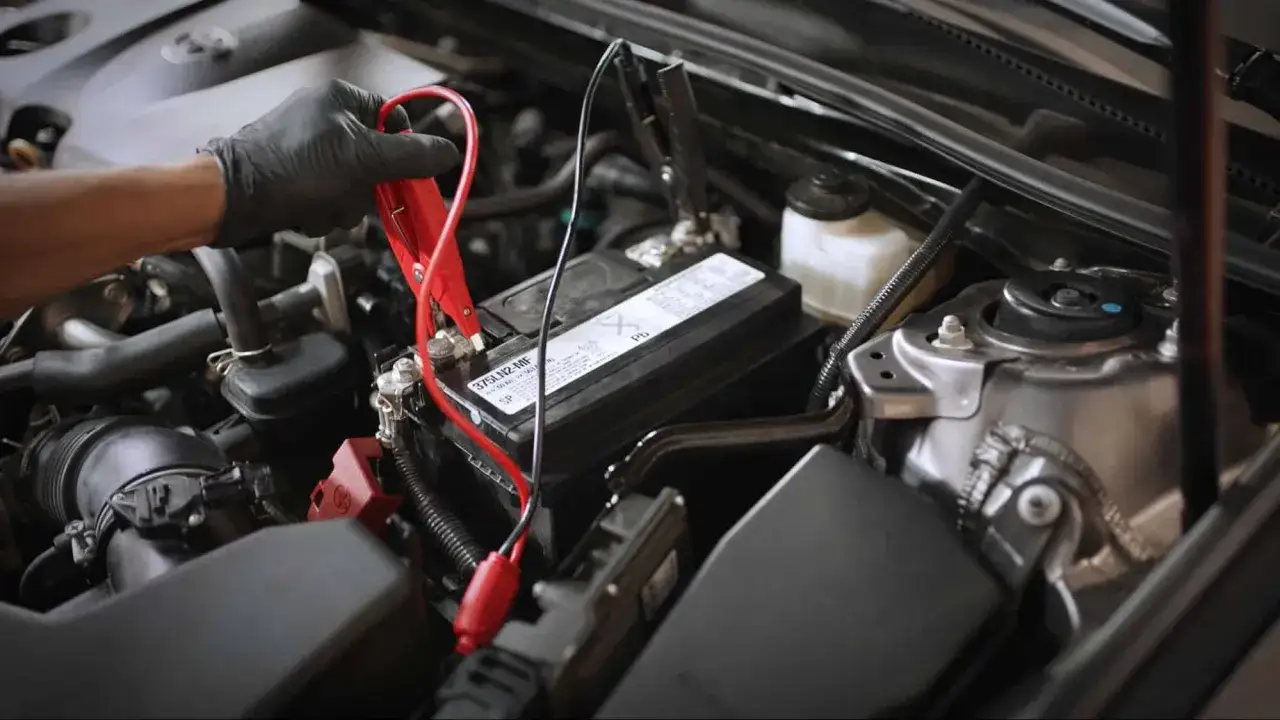 Main d'une personne avec un gant noir connectant des câbles de démarrage à une batterie de voiture. Comment savoir si une batterie ne tient plus la charge.