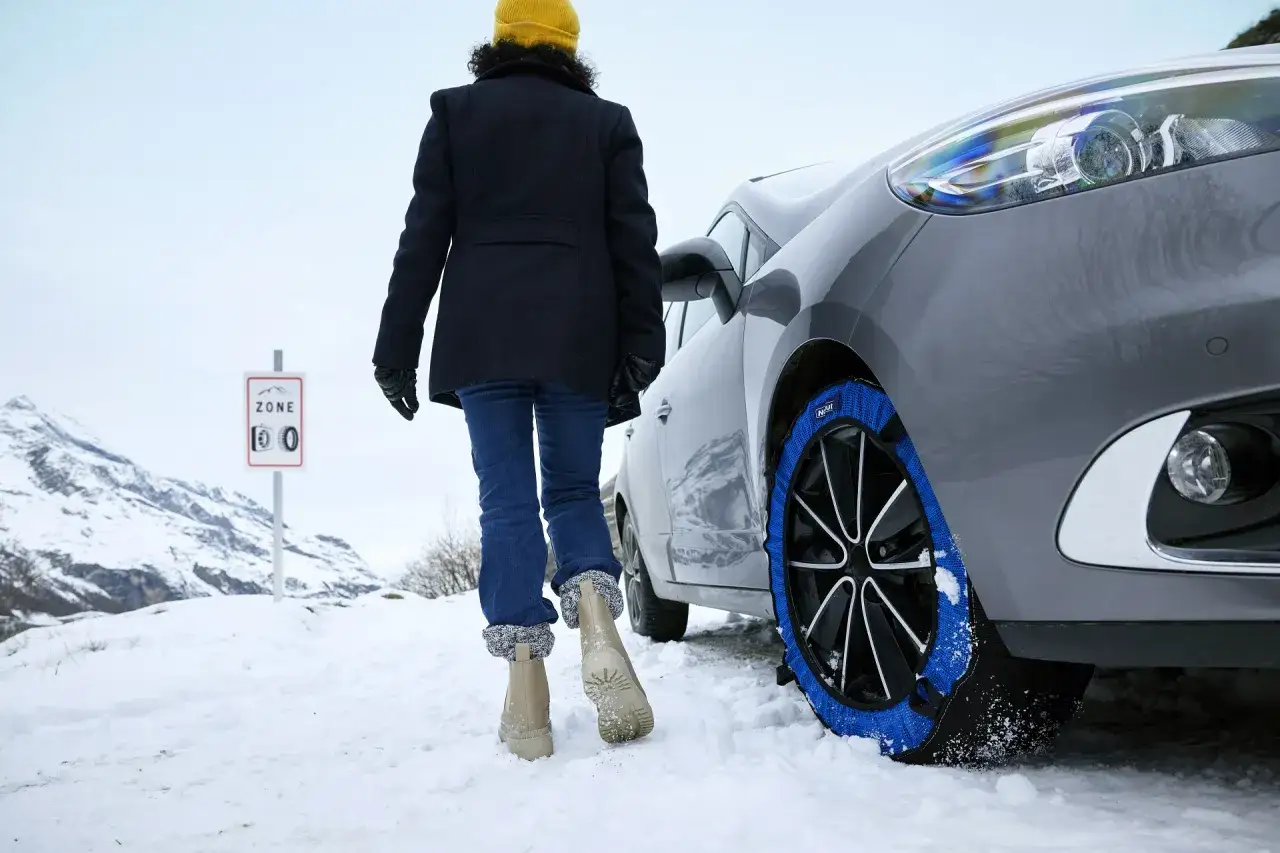 Une personne s'approche de sa voiture équipée de chaînes à neige dans une zone montagneuse. Faut-il enlever la neige sur la voiture ? Le panneau indique une zone où les chaînes sont obligatoires.