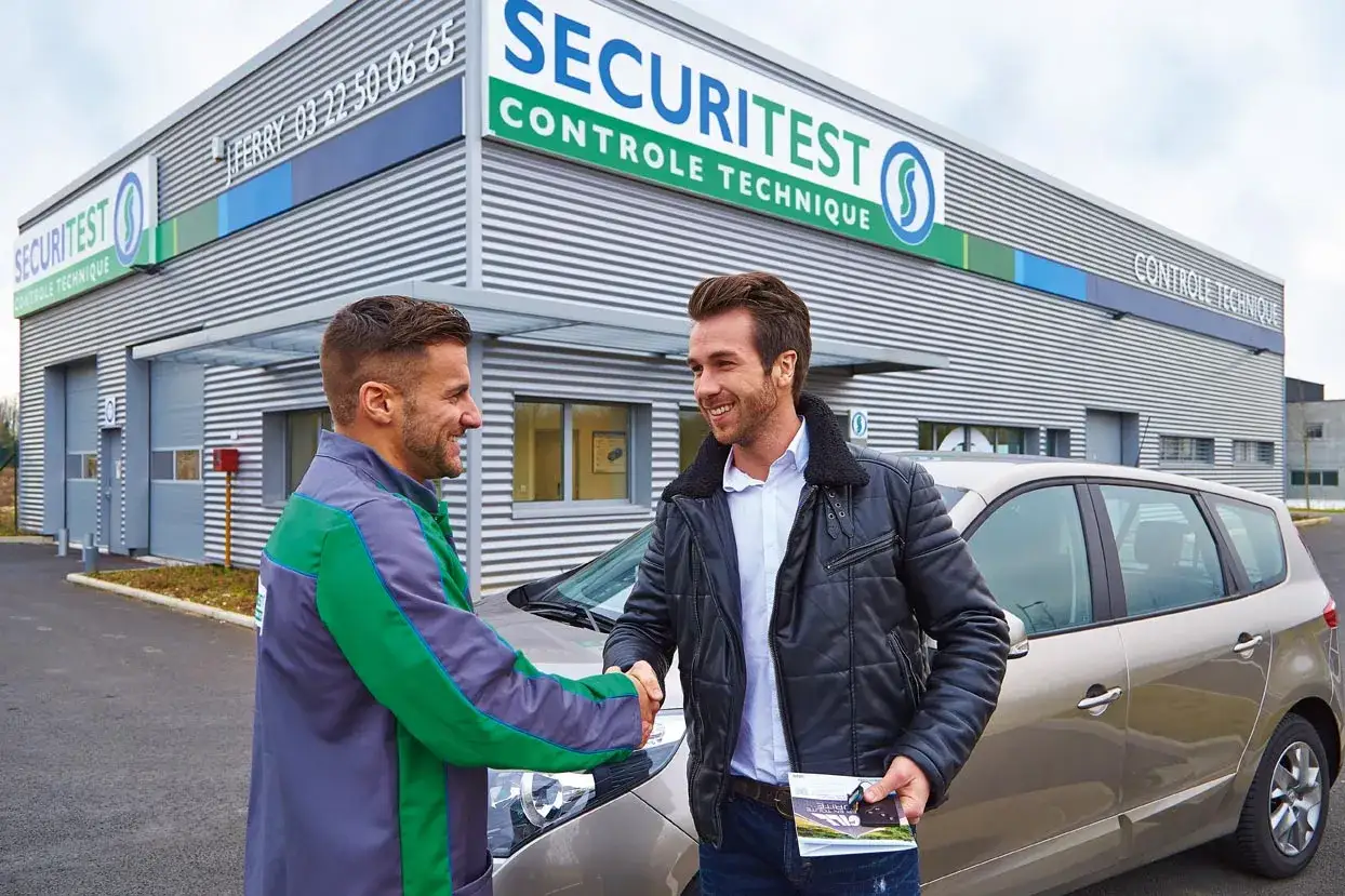 Deux hommes se serrent la main devant un centre de contrôle technique. L'un porte un uniforme vert et gris, l'autre une veste en cuir. Ils discutent peut-être de la ZFE.