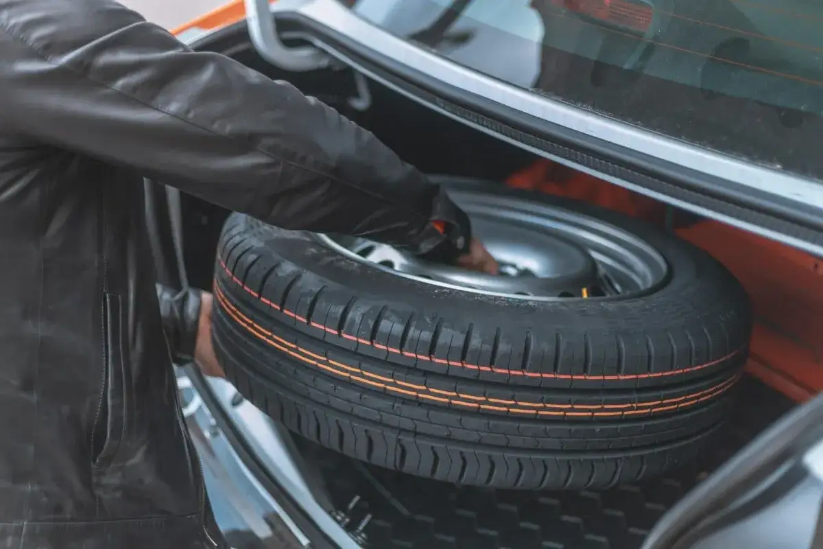 Main dans le coffre, une personne en blouson de cuir sort une roue de secours. Peut-on rouler avec une roue de secours ? Oui, mais avec prudence.