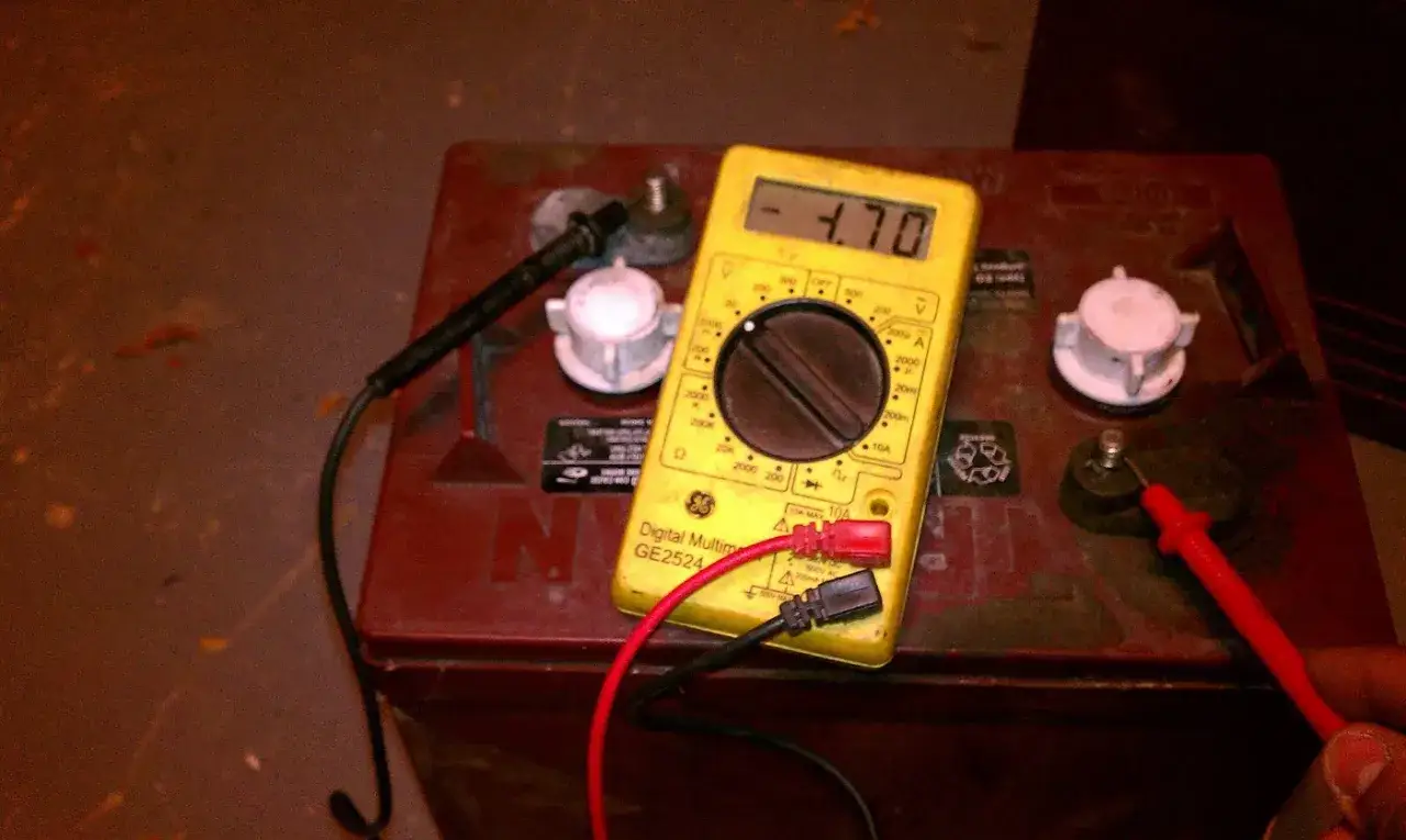 Un multimètre jaune affiche 4.70V sur une batterie de voiture rouge. C'est un indice pour savoir si la batterie de la voiture est morte.
