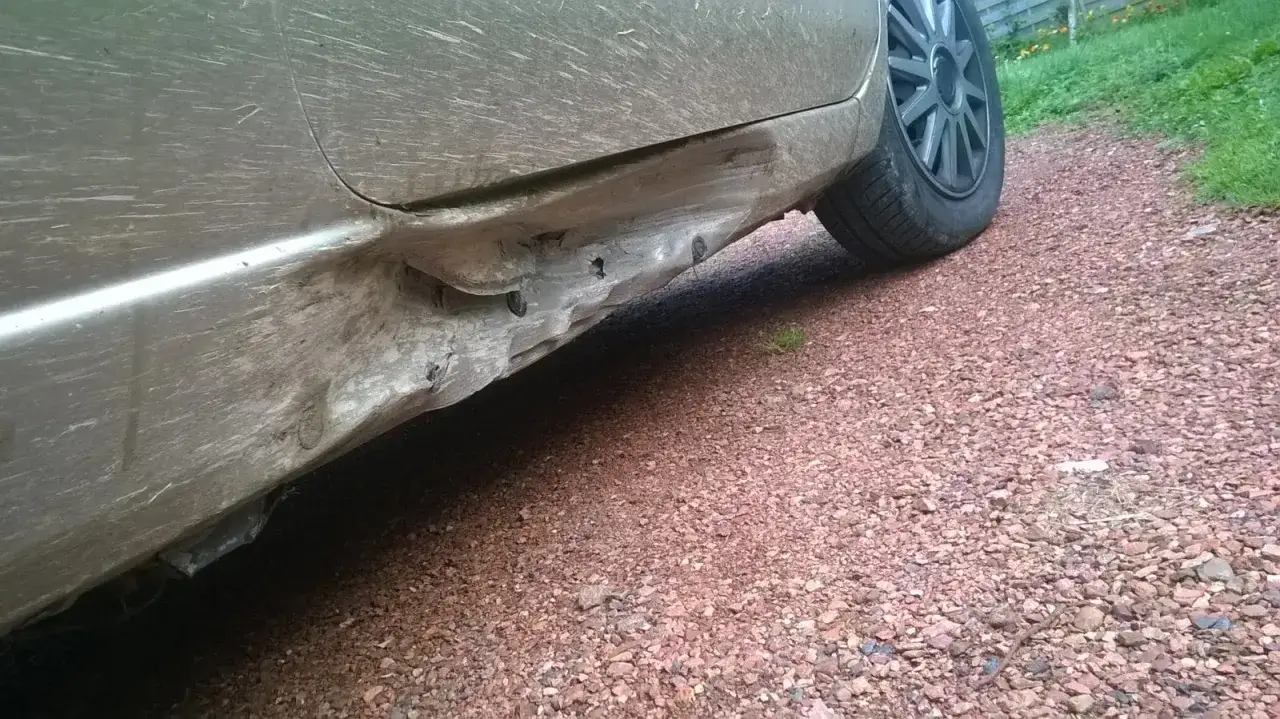 Le bas de caisse de la voiture est gravement endommagé, témoignant d'un choc sous la voiture. La tôle est déformée et arrachée.