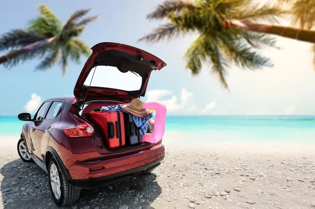 Voiture rouge prête pour le départ en vacances, coffre ouvert rempli de valises, chapeau et bouée rose sur une plage tropicale.