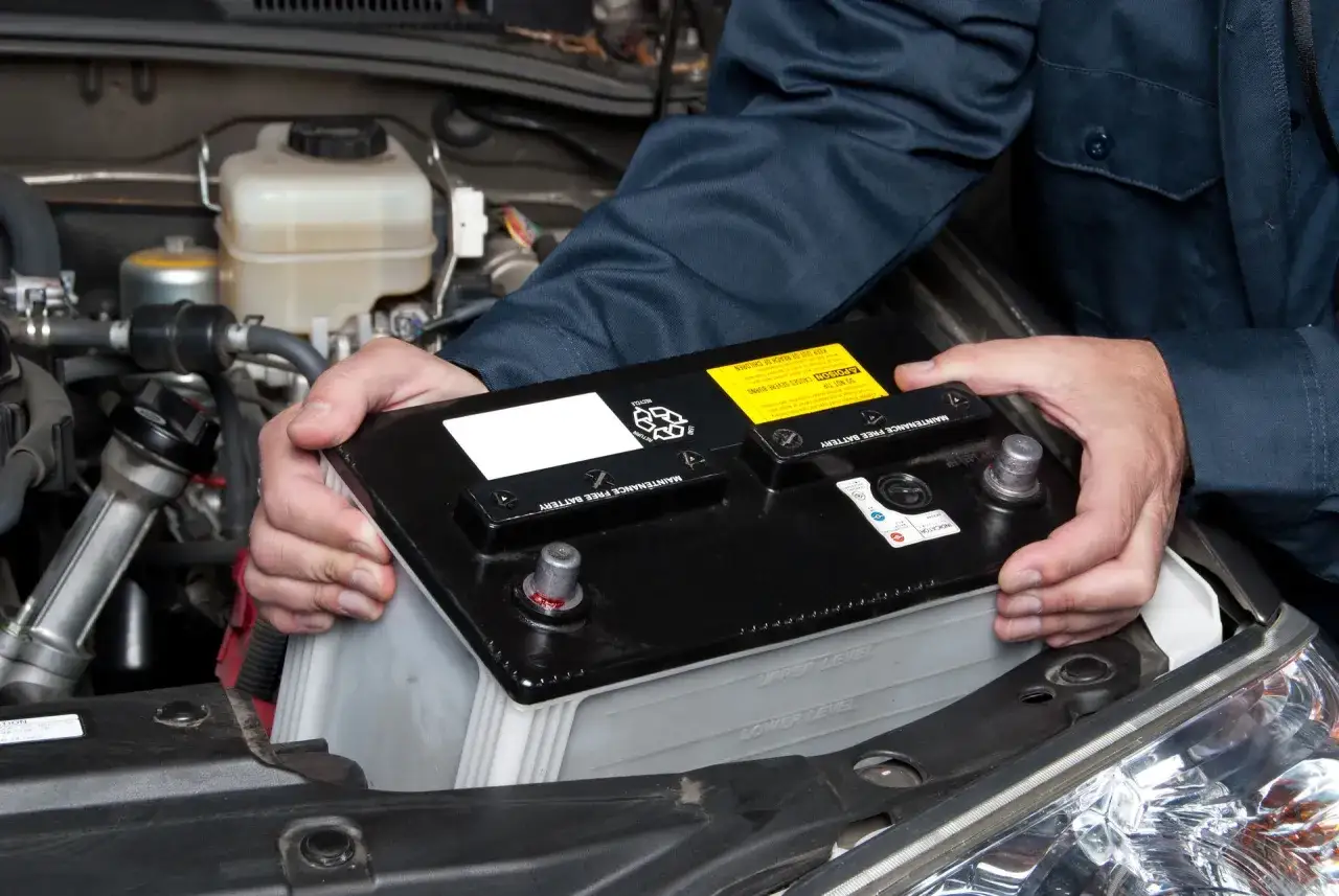 Mains tenant une batterie de voiture pour la changer. Le compartiment moteur est visible.