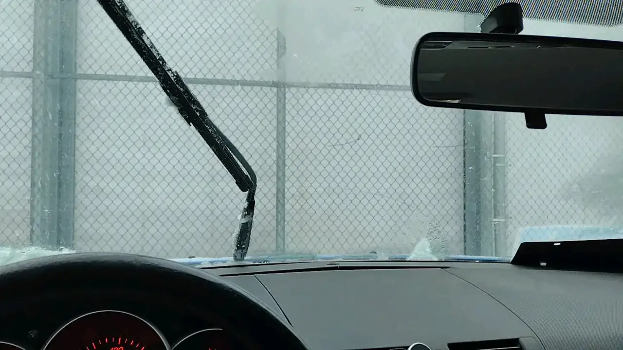 Vue depuis l'intérieur d'une voiture, les essuie-glace qui font du bruit frottent sur un pare-brise embué, devant une clôture en grillage.