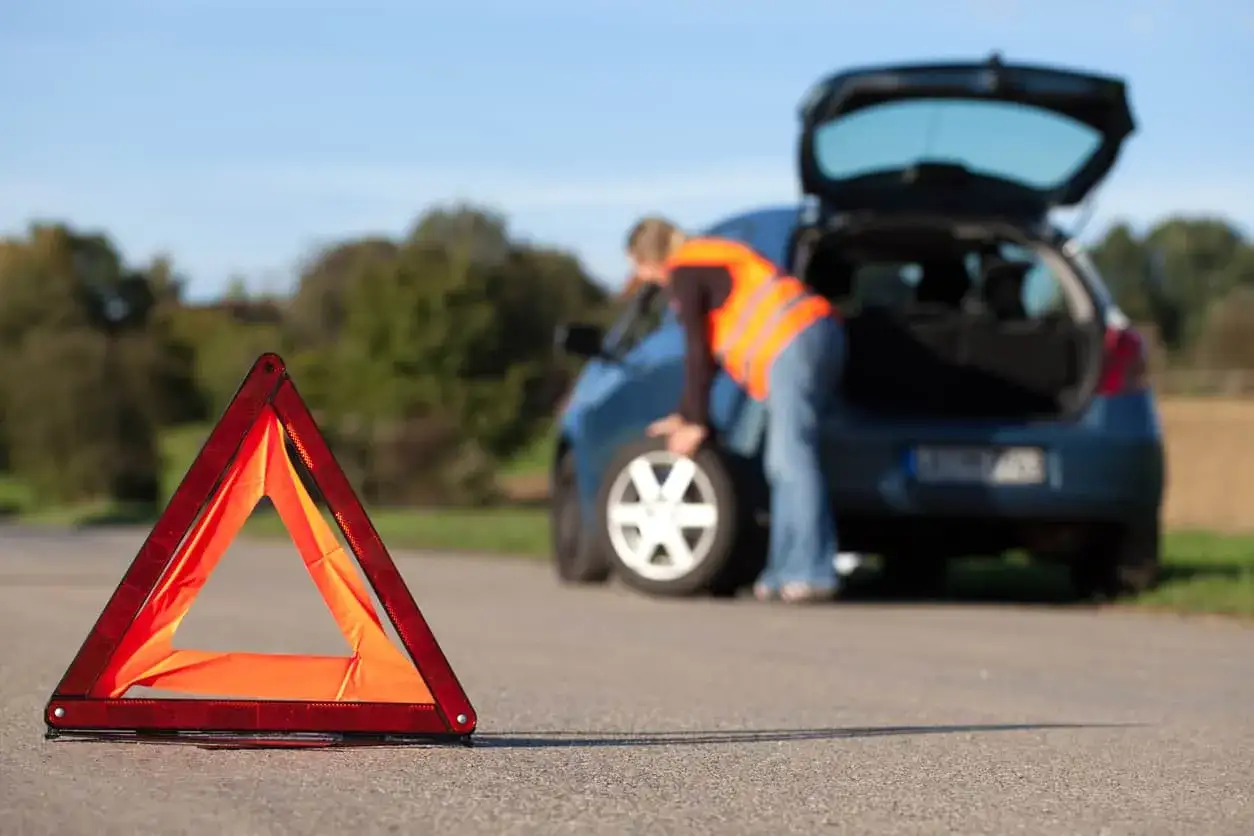 Triangle de signalisation rouge sur la route, une personne en gilet orange s'occupe d'un pneu crevé de sa voiture bleue.