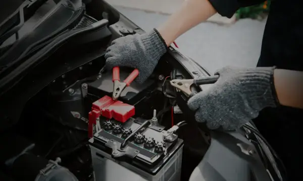 Mains de personne avec des gants gris connectant des câbles à une batterie de voiture. Un symptôme de batterie HS est souvent un démarrage difficile.